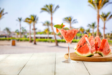 Sticker - Watermelon and a cocktail in glass on wooden tray on white table top. Blur background of sandy tropical beach and palm trees. Empty advertising copy space for products and food.