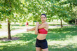 © javiindy - A young woman stretches in a park, wearing red sports bra and black shorts, surrounded by greenery in the sunshine