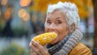 © Lens Legacy - An elderly woman, joyfully eating corn on the cob outdoors, surrounded by warm autumn colors, representing a simple pleasure and the enjoyment of seasonal delights.