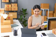 © amnaj - Woman managing online store inventory with tablet and phone, surrounded by boxes, inside a small office space.