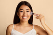 © Prostock-studio - Asian young woman with long black hair applies makeup to her face with a blush brush. She is smiling and looking directly at the camera. She is wearing a white tank top and has a natural makeup look