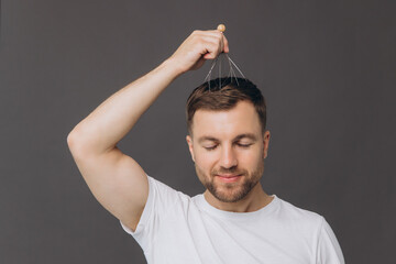 Beauty and health, hair care, a man in a white t-shirt does a head massage with a manual massager on a gray background, head massage equipment