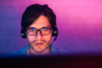 Aufkleber - A young man with dark hair and a beard is wearing glasses and headphones, focused on a computer screen. The room is lit with a purple and pink gradient light, creating a vibrant and modern aesthetic