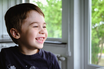 Happy 6 year old boy child is smiling enjoying life. Concept of happy childhood. Portrait of young sitting boy in home background, indoors. Adorable face. Nice smile with beautiful teeth. Side view