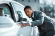 © Prostock-studio - African American man in a suit is inspecting a white car during the day. He has his hand on the car door and is looking at the paint job. The car is parked on a street.
