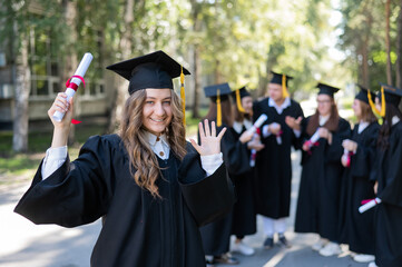 Wall Mural - Group of happy students in graduation gowns outdoors. A young girl with a diploma in her hands in the foreground.