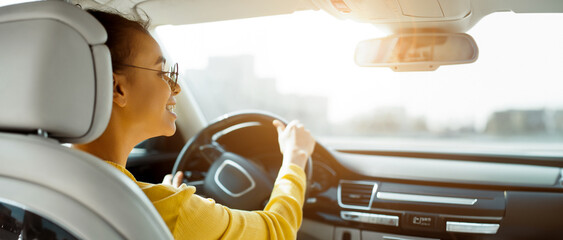 Wall Mural - African American woman in sunglasses smiles while driving a car, with her hand on the steering wheel. The view outside the car window is blurred, with a bright sunny sky in the background.