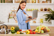 © Prostock-studio - A woman stands in a kitchen holding a laptop, surrounded by fresh produce and a teapot.