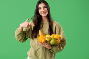 Wall Mural - Pretty young woman pointing at fresh fruits in basket on green background