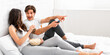 © Prostock-studio - A young couple is comfortably sitting in bed together, watching television and enjoying a bowl of popcorn. The woman is smiling while the man is pointing at the TV and appears to be laughing.