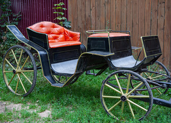 A vintage four-wheeled black horse carriage with red seats parked near a wooden fence on a green grass lawn