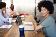 © Wavebreak Media - Smiling at camera, businesswoman with laptop in office, colleagues discussing in background
