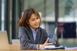 © Songsak C - An Asian businesswoman using a mobile phone smiles and is happy while sitting at a working desk in a modern office.