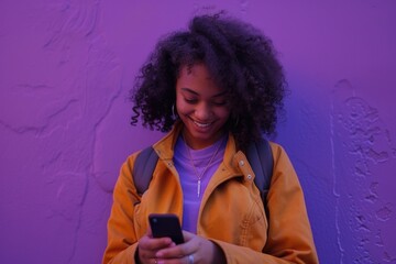 Wall Mural - A woman with curly hair is smiling while looking at her cell phone. She is wearing a yellow jacket and a purple shirt