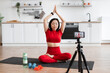 © sofiko14 - Woman in red sportswear meditating on yoga mat in kitchen. Online fitness session with camera on tripod. Healthy lifestyle, mindfulness, home workout.