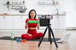 © sofiko14 - Woman in red activewear sitting on yoga mat using resistance bands while filming workout video. Home exercise equipment, water bottle, and laptop nearby.