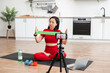 © sofiko14 - Woman in red sportswear demonstrating exercises with resistance band during online fitness class. Filming with smartphone, laptop, water bottle, and dumbbells visible. Modern kitchen background.