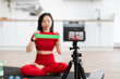 © sofiko14 - Woman in red activewear recording yoga fitness tutorial at home using smartphone. She is showing resistance band exercises sitting on yoga mat in modern kitchen.