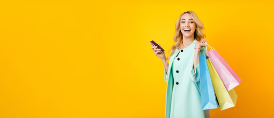 A young woman with blonde hair is smiling at the camera while holding colorful shopping bags in one hand and a phone in the other. She is wearing a light blue coat, panorama with copy space
