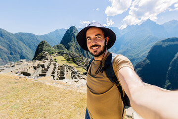 Happy young traveler man taking selfie portrait while enjoying vacation in Peru. Joyful tourist visiting Machu Picchu. Travel and holiday lifestyle concept.