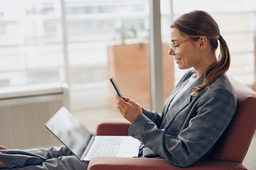 Wall Mural - Stylish female freelancer in eyeglasses working on laptop and use phone while sitting in coworking 