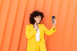 © VISTA by Westend61 - Smiling beautiful woman wearing yellow blazer using smart phone standing in front of orange wall