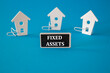 © Liudmila - Fixed assets symbol. Wooden houses sits next to a wooden black board with the word fixed assets. Beautiful blue background. Business and fixed assets concept.