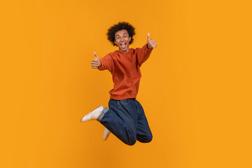 African American young man with an afro is jumping in the air with both thumbs up. He is wearing a red sweater and blue jeans. He is smiling and looking directly at the camera