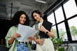 © Bliss - Two women stand in a modern office, reviewing documents together.