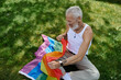 © Bliss - A mature gay man with tattoos and a grey beard sits on the grass holding a rainbow flag.