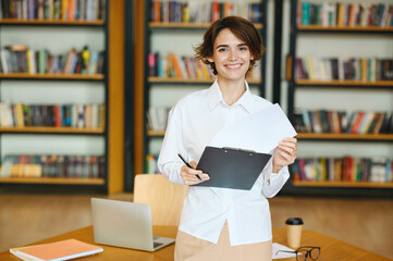 Wall Mural - Young fun successful employee business woman wear white shirt casual clothes hold clipboard with paper account documents stand near table work at office desk with pc laptop Achievement career concept