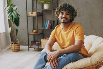 Wall Mural - Young smiling happy cheerful fun Indian man he wearing orange casual clothes look camera sits in armchair stay at home hotel flat rest relax spend free spare time in living room indoor Lounge concept