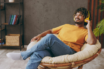 Wall Mural - Young happy Indian man wear orange casual clothes listen to music in headphones sits in armchair couch stay at home hotel flat rest relax spend free spare time in living room indoor. Lounge concept.