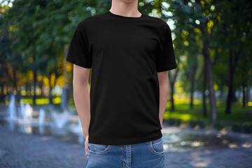 Wall Mural - Mockup of a casual black cotton t-shirt on a guy with hands in pockets, on the background of a park, fountain, trees, front view.