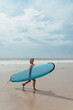 © blackday - Cheerful woman carries a blue surfboard while walking on the beach