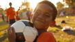 © JR-50 - Happy young boy smiling and holding a soccer ball with teammates in the background on a sunny day at the park.