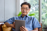 Fototapeta Zachód słońca - Senior Asian man using tablet connect internet while sitting on sofa at home