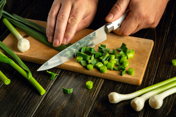 Wall Mural - A cook slices young green garlic stalks with a knife on a wooden cutting board to prepare dinner.