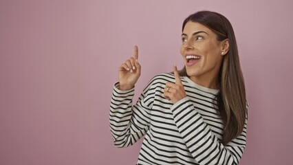 Sticker - Attractive young hispanic woman in striped t-shirt, stands confidently over pink isolated background. with a cheerful smile, she points to the side, inviting the camera's attention.