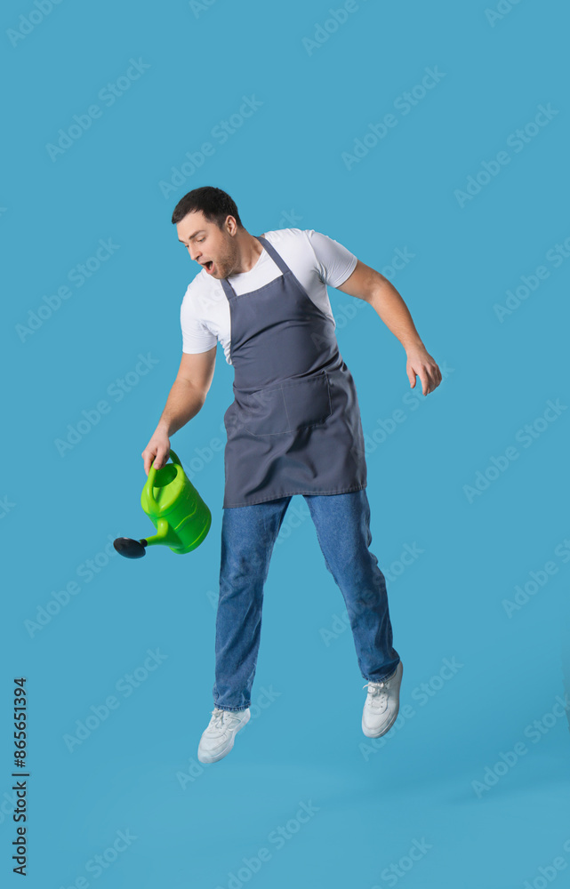 Young male gardener jumping with watering can on blue background