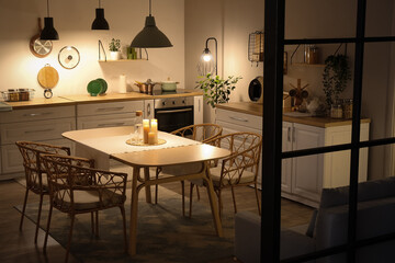 Wall Mural - Interior of modern kitchen with glowing lamps, white counters, sofa and dining table at evening