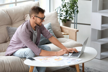 Poster - Young businessman in eyeglasses working with laptop and documents on sofa in office