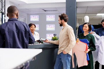 Wall Mural - Man standing in line making contactless smartphone payment, paying for purchases with electronic credit card while shopping on Black Friday. Diverse people holding clothes waiting at cash regiter desk