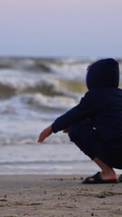 Wall Mural - Boy resting near the sea at sunset. Child is painting on sandy beach on foamy waves background of the evening sea. Vertical video