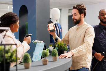 Wall Mural - Man customer holding smartphone showing mobile discount code to saleswoman while shopping in clothing store. Cashier scanning promo coupon while serving shopper at checkout counter