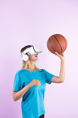 Wall Mural - Girl wearing visual reality glasses and casual cloth and holding basketball. Caucasian woman playing basketball while standing at pink background at sport arena hologram. Innovation. Contraption.