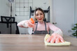 © Natee Meepian - Professional Maid Cleaning a Modern Home Office with a Smile, Wearing Protective Gloves and Apron, Ensuring a Spotless