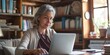 © Iryna - Senior businesswoman with grey hair, sitting at desk, using laptop, enjoying online work at home.