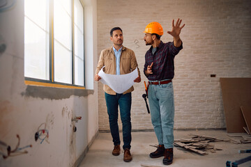 Home owner and manual worker talking while analyzing blueprints at construction site.