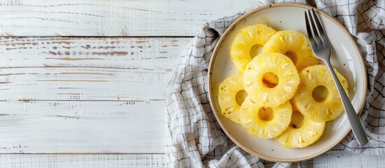 Poster - Top view of a plate displaying canned pineapple rings accompanied by a fork, kitchen towel, on a white wooden table, with copy space image available.